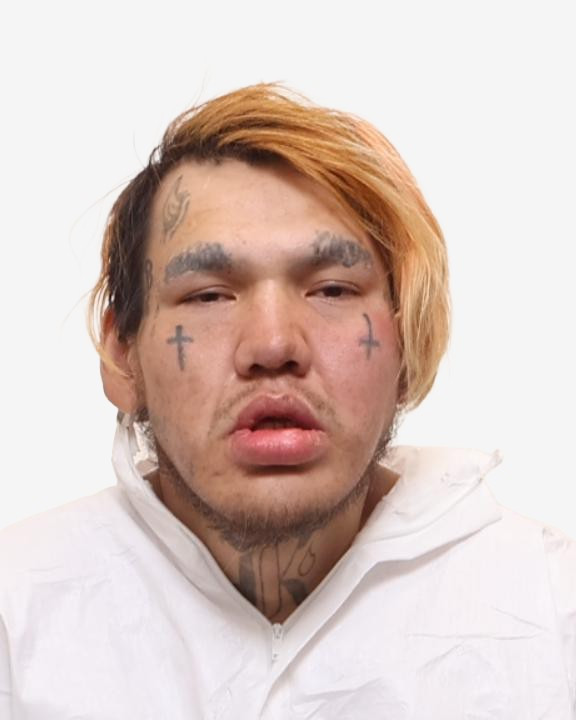 Close-up photo of a man with two-tone hair, facial tattoos including crosses on the cheeks, and a neck tattoo, wearing a white hooded garment against a plain white background.