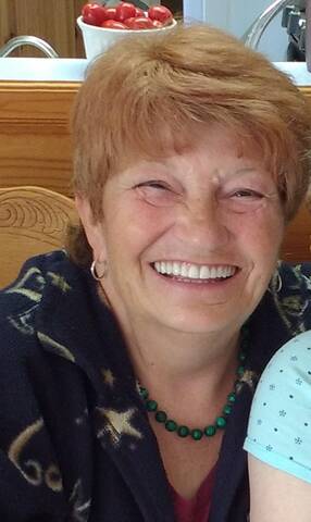 Smiling woman with short reddish hair, wearing a green beaded necklace and a dark top with star patterns, seated indoors near a kitchen counter.