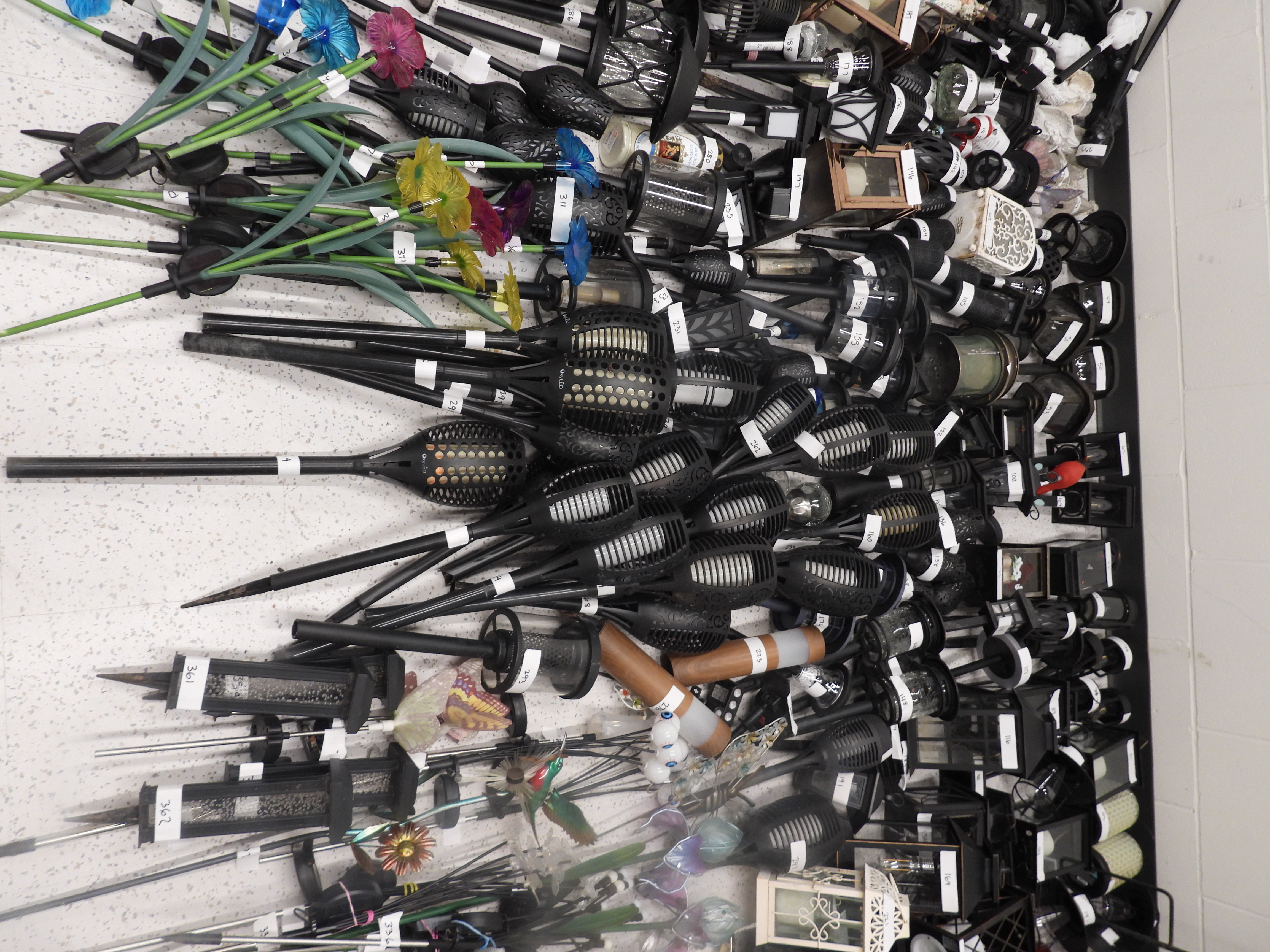 Many seized solar garden lanterns, torches, glass flower stakes and decorative items laid out on a wall, each tagged with numbered labels.