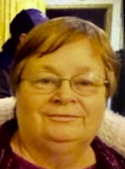 Close-up photo of an older woman with short reddish-brown hair, wearing glasses and a dark top with a light collar, smiling slightly, with a blurred background.