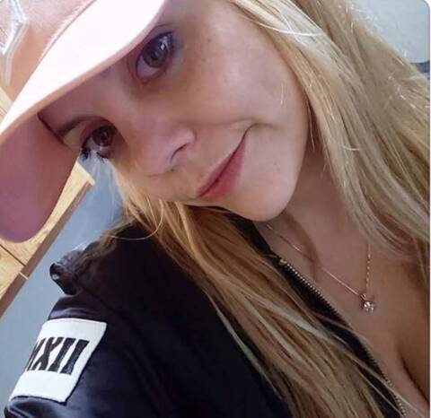 Selfie-style photo of a smiling woman with long blonde hair, wearing a pink cap and a dark zippered jacket with a patch, and a thin necklace. The page text labels it as a photo of the person named in the appeal.