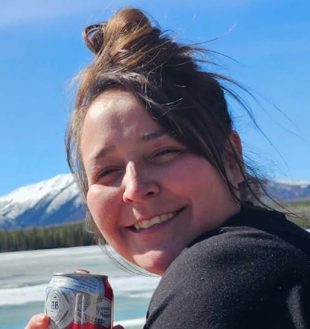 Smiling woman with brown hair pulled up in a bun, holding a can outdoors, with snowy mountains and a frozen lake behind her.