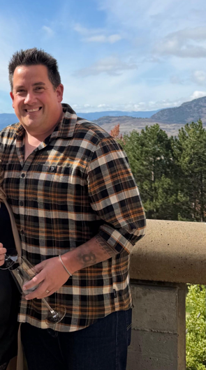 A smiling man in a plaid flannel shirt standing outdoors on a stone ledge with hills and trees behind him.