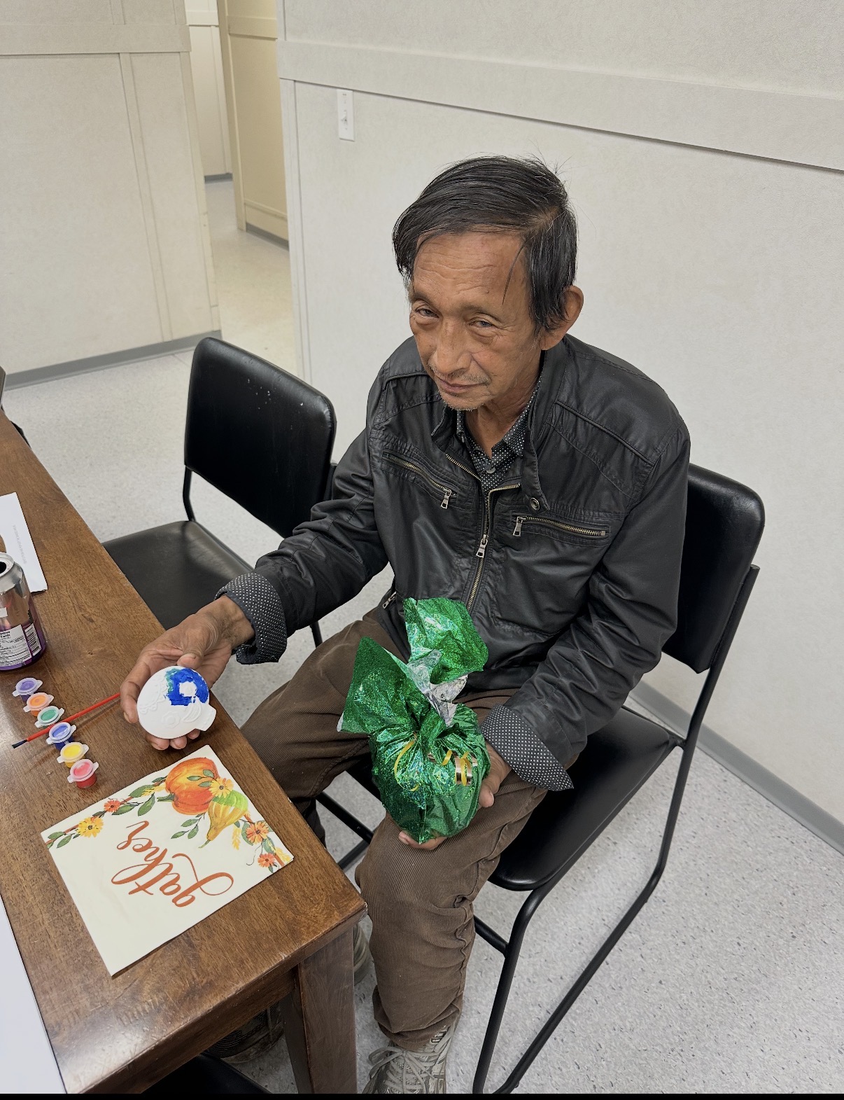 An older man seated indoors at a table with paint pots and a painted ceramic object, holding a green wrapped gift, wearing a dark jacket.