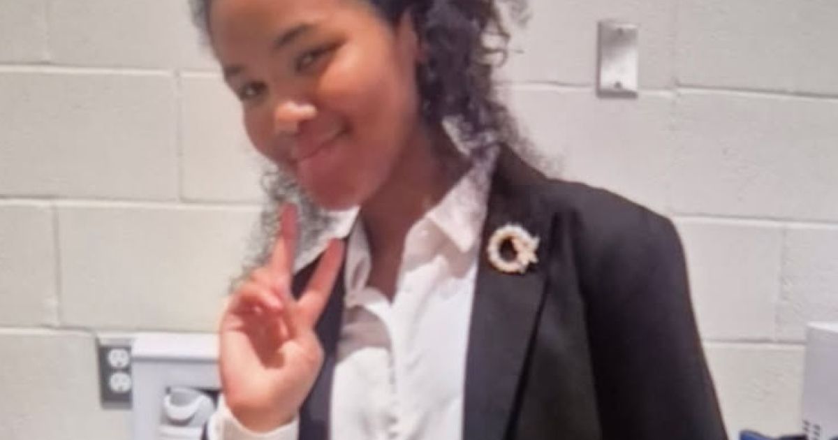 A smiling young person with curly dark hair in a white collared shirt and dark blazer with a small circular pin, making a peace sign against a white cinder block wall.