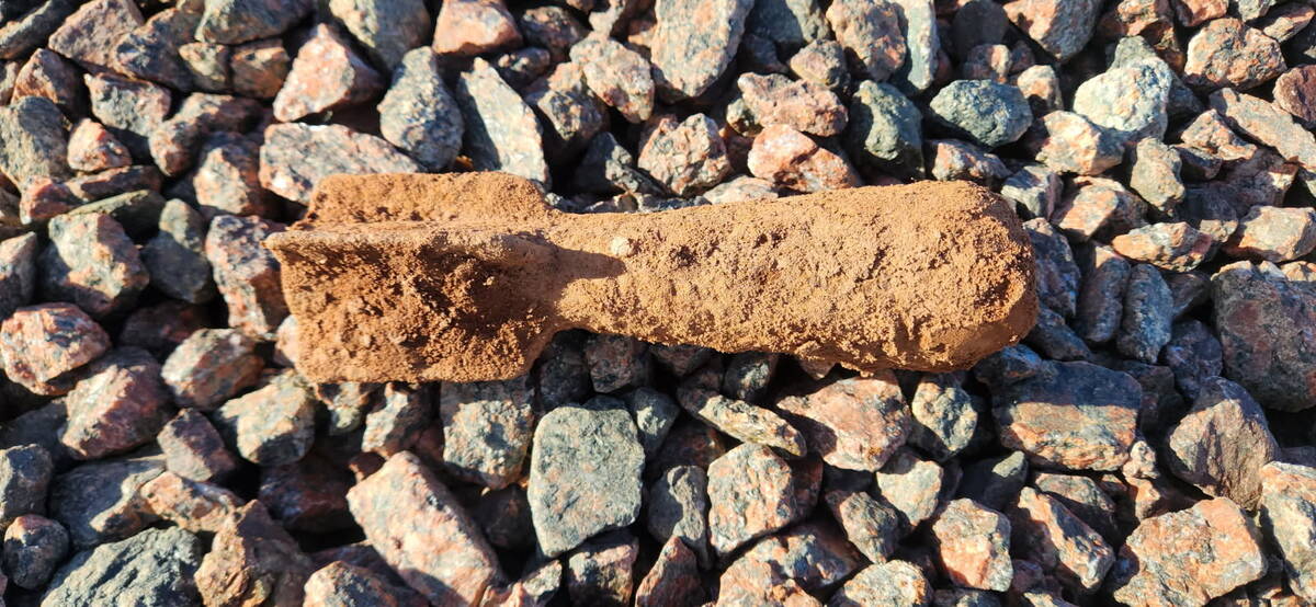 A rusty, dirt-covered metal cylinder lying on a bed of reddish-brown gravel stones, consistent with the possible explosive ordnance found in the PEI field.