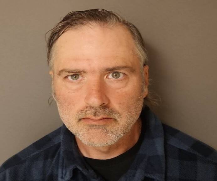 Head-and-shoulders photo of a man with greyish swept-back hair and stubble, wearing a dark blue plaid shirt against a plain grey background.