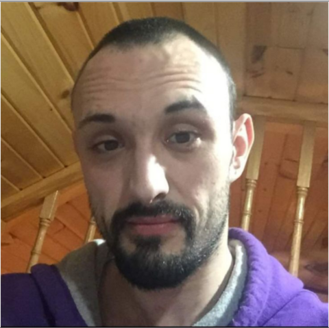A man with short dark hair and a dark beard, wearing a purple hooded sweatshirt, photographed in front of a wood-paneled ceiling and wooden railing.