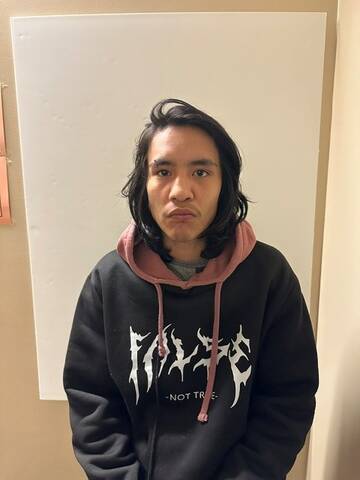 Photo of a young man with shoulder-length dark hair wearing a black hoodie with a pink hood and white graphic lettering, standing against a white wall.