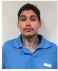 Booking-style photo of a man with short dark hair and a mustache, wearing a blue polo shirt, with visible tattoos on his neck and chest against a plain light background.